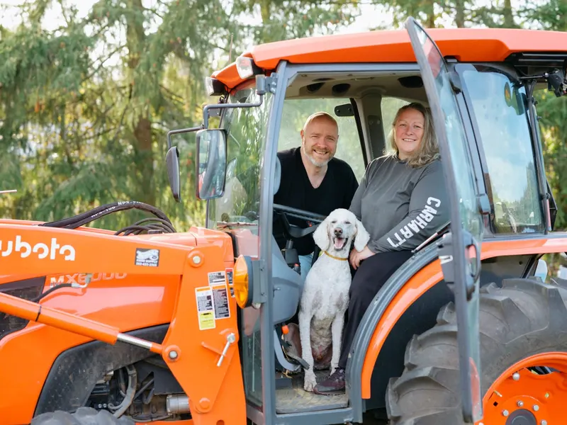 Andrew and his sister Lindsay smiling in the cab of an orange Kubota tractor with Ozzy, a white standard poodle