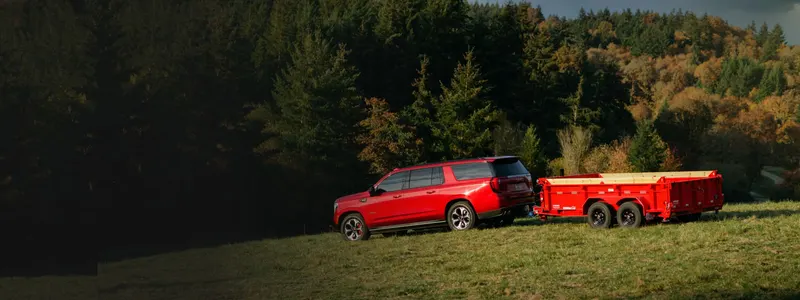 A red SUV towing a red Red Wagon Rentals dump trailer across a green field in front of an Oregon forest